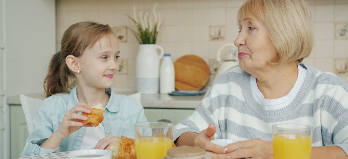 Kid adorable girl is eating croissant and talking to happy granny laughing having breakfast at kitchen table. Family relationship and childhood concept.