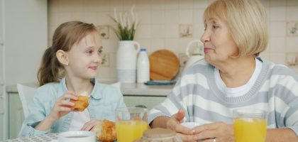Kid adorable girl is eating croissant and talking to happy granny laughing having breakfast at kitchen table. Family relationship and childhood concept.
