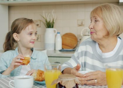 Kid adorable girl is eating croissant and talking to happy granny laughing having breakfast at kitchen table. Family relationship and childhood concept.